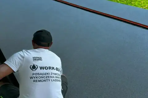 Pracownik w koszulce z napisem "POSADZKI ŻYWICZNE" wykonuje prace na gładkiej, ciemnej powierzchni.