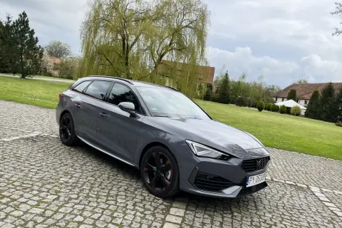Szary Cupra kombi na brukowanym podjeździe, z zielonym trawnikiem i drzewami w tle.
