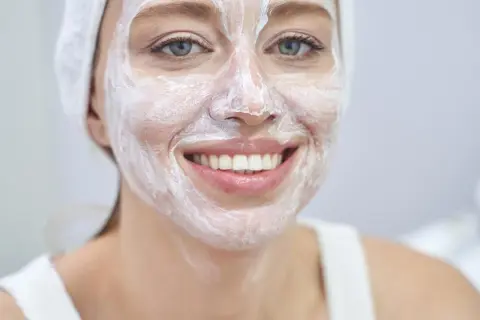 Lächelnde Frau mit weißer Gesichtsmaske und Handtuch auf dem Kopf. Wie oft Maske Gesicht? Regelmäßige Pflege für strahlende Haut.