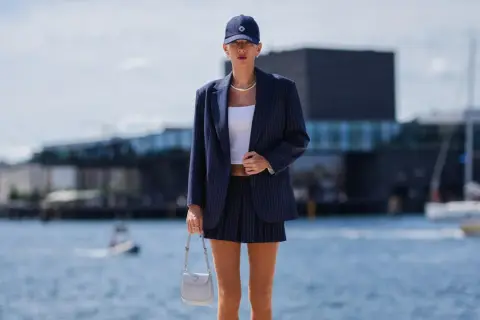 Ein gestreifter, **blauer Blazer kombiniert** mit einem kurzen Rock und weißem Top. Eine Frau trägt das Outfit am Wasser.
