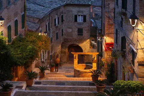 Un vicolo illuminato a San Giorgio di Pesaro, con un pozzo antico e una persona che sale le scale.