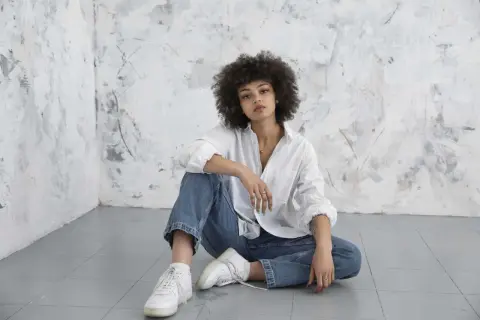 Kobieta z kręconymi włosami w białej koszuli i jeansach, siedząca na podłodze. Jej styl to klasyczny, basic look.