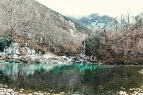 El río turquesa fluye sereno, reflejando árboles desnudos y montañas neblinosas. Un paisaje que evoca la paz de la olla de San Vicente.