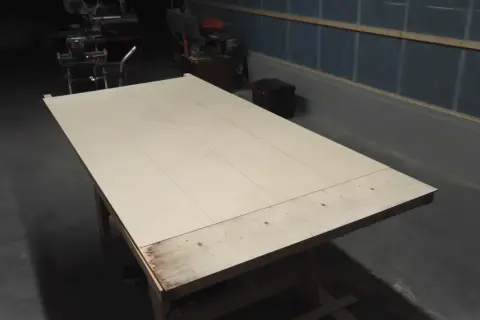 Stor skiva av plywood på ett arbetsbord, redo att bygga slagport. Verktyg syns i bakgrunden.