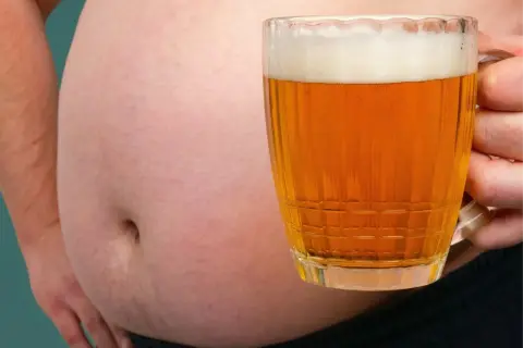 Ein Bierbauch ist da, weil Bier dick macht. Ein Mann hält ein Glas Bier vor seinem Bauch.