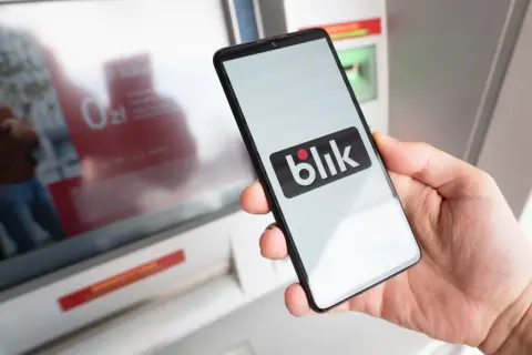 Dłoń trzyma smartfon z logo Blik. Obok bankomat, który ułatwia szybkie płatności. Kto wymyślił Blik? To polski system płatności.