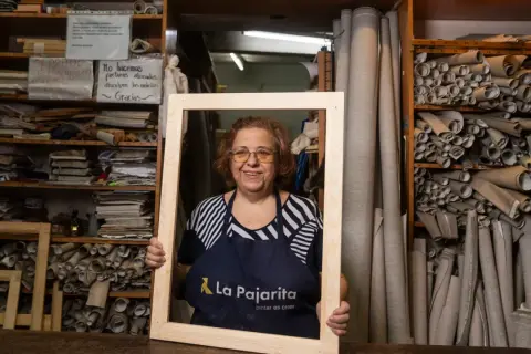 Mujer sonriente en taller, rodeada de rollos de tela y estanterías con materiales. Sostiene un marco de madera en rollo.