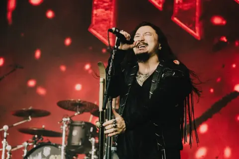 Wokalista zespołu rockowego na scenie, z mikrofonem w dłoni, w tle czerwone światła i perkusja. Koncert w Hali Stulecia we Wrocławiu.