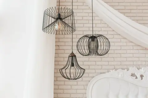 Nowoczesne **lampa pająk aranżacje** w loftowym stylu. Trzy wiszące lampy z drucianymi kloszami na tle ceglanej ściany i białej zasłony.