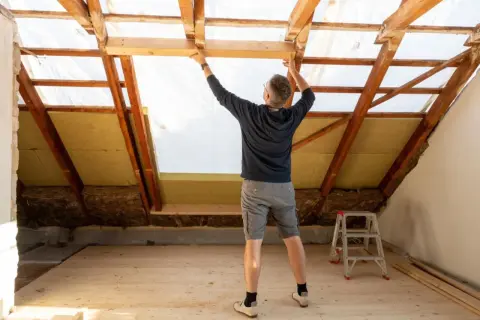 Mężczyzna montuje drewnianą konstrukcję poddasza, gdzie widać już wełnę mineralną. Zastanawiasz się, ile kosztuje metr ocieplenia poddasza?