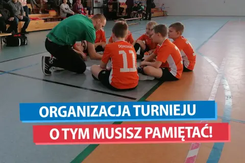 Trener w zielonej koszulce tłumaczy dzieciom w pomarańczowych strojach, jak zorganizować turniej piłki nożnej.