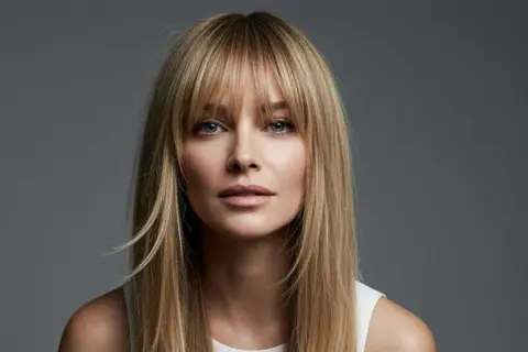 Blonde Frau mit Curtain Bangs, die ihr Gesicht umrahmen. Ihr Blick ist direkt in die Kamera gerichtet.