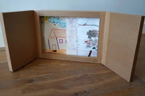 Un kamishibai casero muestra dibujos infantiles: una casa, un paisaje rural y un pueblo con un tren.