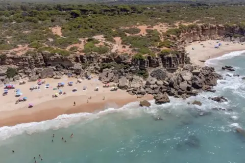 Una cala enebro con acantilados rocosos, arena dorada y aguas turquesas. Gente disfrutando del sol y el mar bajo sombrillas.
