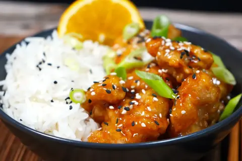 En skål med ris och krispig, glaserad orange chicken, toppad med sesamfrön och salladslök. En apelsinskiva vilar bakom.