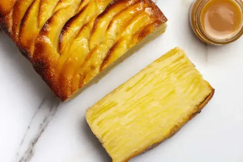 Une tranche de tarte aux pommes, dorée et caramélisée, à côté d'une autre tranche montrant la texture fine des pommes. Un petit pot de caramel est aussi visible.
