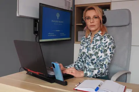 Kobieta w okularach i słuchawkach siedzi przy biurku, pracuje na laptopie. Na monitorze widnieje logo i dane kontaktowe.