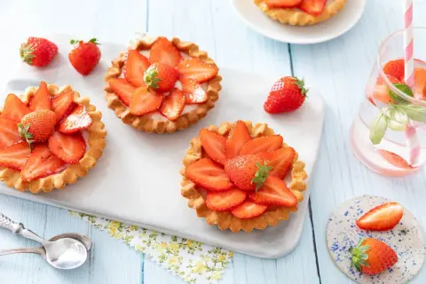 Trois tartelettes aux fraises, garnies de crème et de fruits rouges frais, sur un plateau blanc. Une fraise entière repose à côté.