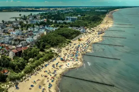 Tłum ludzi na plaży w Mielnie, cieszących się słońcem i morzem. W tle widać zabudowania i jezioro. To idealne miejsce na wakacje, pełne atrakcji.