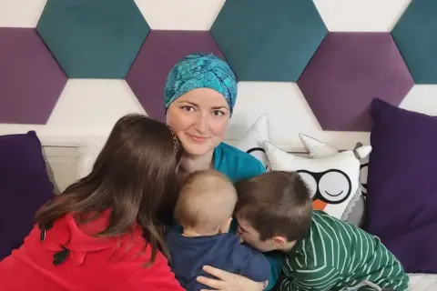 Mama w chustce i trójka dzieci. Najstarszy syn całuje maluszka, córka przytula się do mamy. W tle heksagony i poduszki. BIRADS 4a.