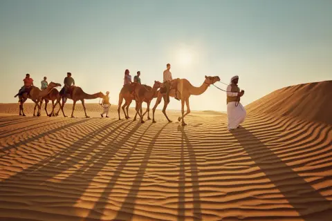 Kamelkarawane in der Wüste bei Sonnenuntergang. Ein Führer in traditioneller Kleidung begleitet Touristen. Tolle Abu Dhabi Tipps für ein unvergessliches Erlebnis.