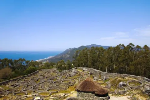 Ruinas circulares de piedra en el monte Santa Tecla, con el mar y montañas al fondo.
