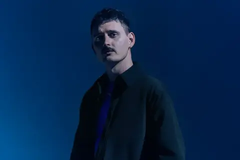 Dawid Podsiadło w ciemnej kurtce i fioletowej koszulce na niebieskim tle.