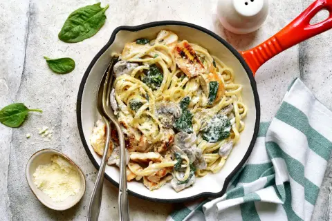 Kremowy makaron z kurczakiem, szpinakiem i grzybami w patelni, posypany parmezanem.