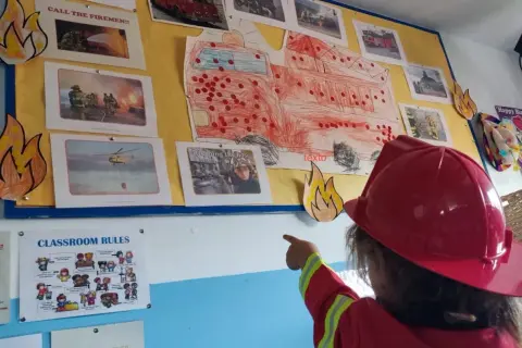 Niña con casco de bombero señala un mural de bomberos, parte de originales proyectos infantiles.