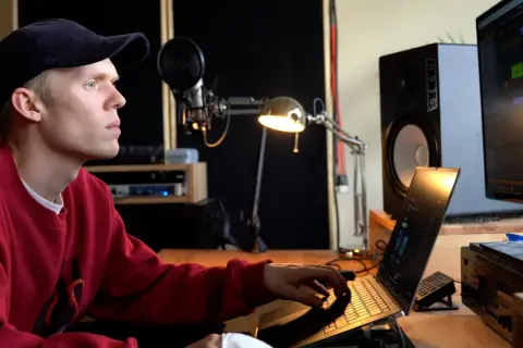Ein junger Mann mit rotem Pullover und Kappe arbeitet an einem Laptop in einem Musikstudio. Er komponiert Musik, die die **lofi bedeutung** von Entspannung und Kreativität verkörpert.