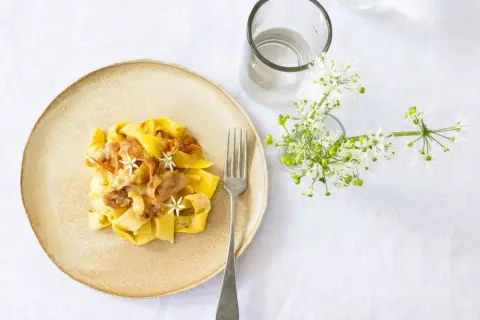 Ein Teller mit cremiger Pasta, die wie Fettuccine aussieht, garniert mit zarten Blüten und Speck. Daneben ein Glas Wasser und ein Strauß weißer Blumen.
