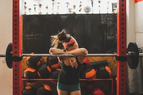 Erschöpfte Athletin lehnt an Hantelstange. Eine Woche Trainingspause ist sinnvoll für Regeneration und neue Motivation.