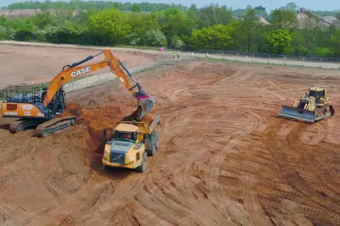 Koparka i wywrotka pracują na placu budowy, niwelując teren. W tle spychacz. Kary za niwelację terenu mogą być wysokie.