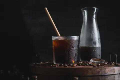 Ett glas med kallpressat kaffe, isbitar och en träpinne på en träbricka med kaffebönor. En karaff med kaffe står bredvid.