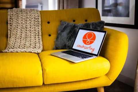 Żółta kanapa z laptopem otwartym na stronie couchsurfing. Obok koc i szara poduszka.
