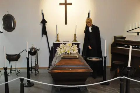 En präst med sorgflor på hatten står bredvid en kista med blommor. En orgel och kors syns i bakgrunden.