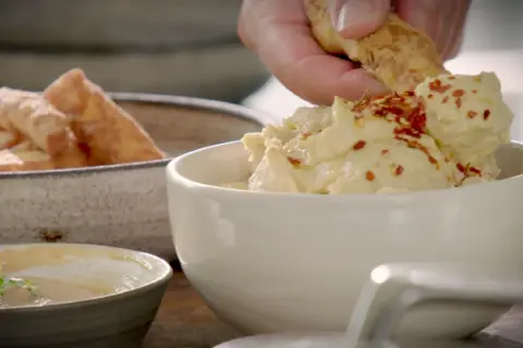 En hand doppar en bit i krämig hummus toppad med chiliflakes. Bredvid står en skål med gyllene halloumi fries.