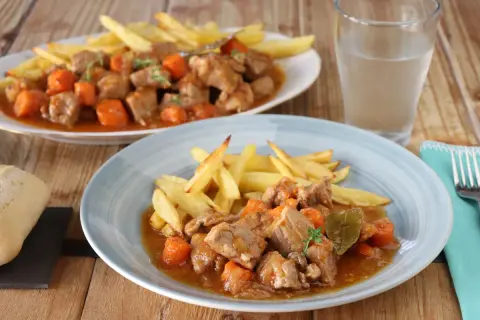 Dos platos de estofado de cerdo con patatas fritas y zanahorias, servidos en una mesa de madera.