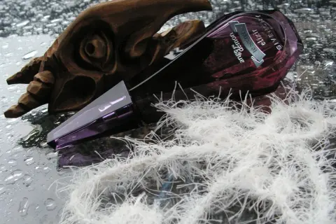 Givenchy Ange ou Demon Le Secret Elixir w fioletowej butelce, obok drewnianej rzeźby i białych włókien.