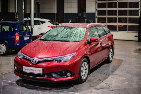 Czerwona Toyota Auris 1.6 benzyna z kroplami deszczu na karoserii, zaparkowana w garażu obok innych samochodów.