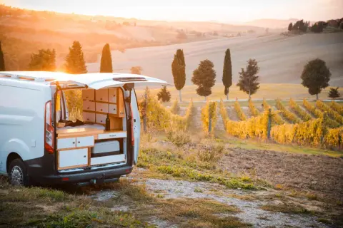 Un camper in un vigneto al tramonto, con dolci colline toscane sullo sfondo. L'interno del camper è visibile, con un letto e mobili.