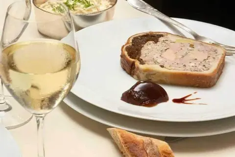 Une tranche de pâté en croûte, garnie de confiture de figues, accompagnée d'un verre de vin blanc et d'une coupelle de crudités.