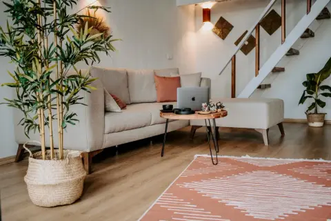 Przytulne, małe mieszkanie w stylu boho. Jasna kanapa, drewniany stolik z laptopem i ozdobami, bambus w koszu, schody i rośliny tworzą ciepłą atmosferę.