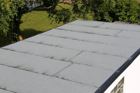 Graues Garagendach mit sichtbaren Rissen und Spalten, die auf ein undichtes Garagendach hindeuten. Im Hintergrund grüner Rasen und Bäume.