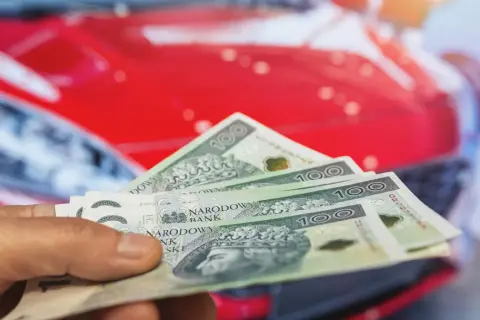 Dłoń trzyma banknoty 100 zł na tle czerwonego samochodu. Koszty posiadania auta, w tym podatek od samochodu, mogą być znaczące.