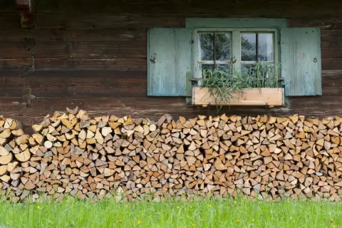 Ein ordentlicher Stapel Holz liegt vor der rustikalen Hauswand. Ein Fenster mit blauen Fensterläden und einem Blumenkasten mit Kräutern darüber.