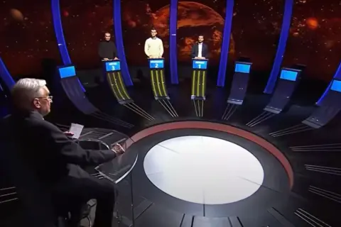 Trzech uczestników teleturnieju stoi przy swoich stanowiskach, a prowadzący siedzi przodem do nich.