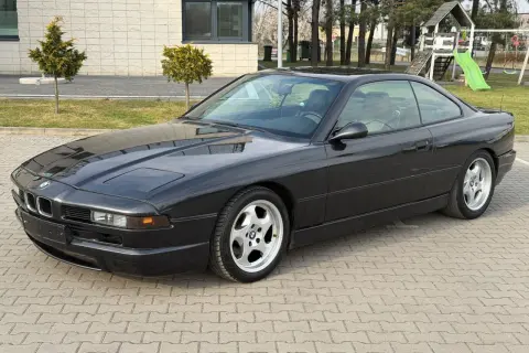Czarne BMW V12, klasyk z lat 90., zaparkowane na kostce brukowej przed budynkiem z placem zabaw w tle.