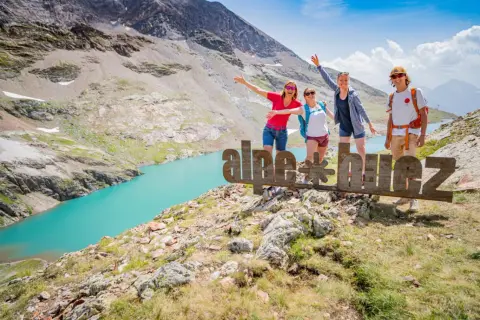 Quatre amis profitent de leurs vacances d'été dans les Alpes, près d'un lac turquoise. Ils posent devant un panneau "alpe d'huez".