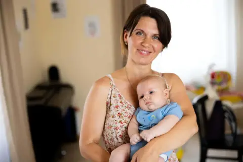 A smiling mother holds her 2-month-old baby, celebrating precious milestones. The baby wears a blue onesie with a yellow and blue heart.